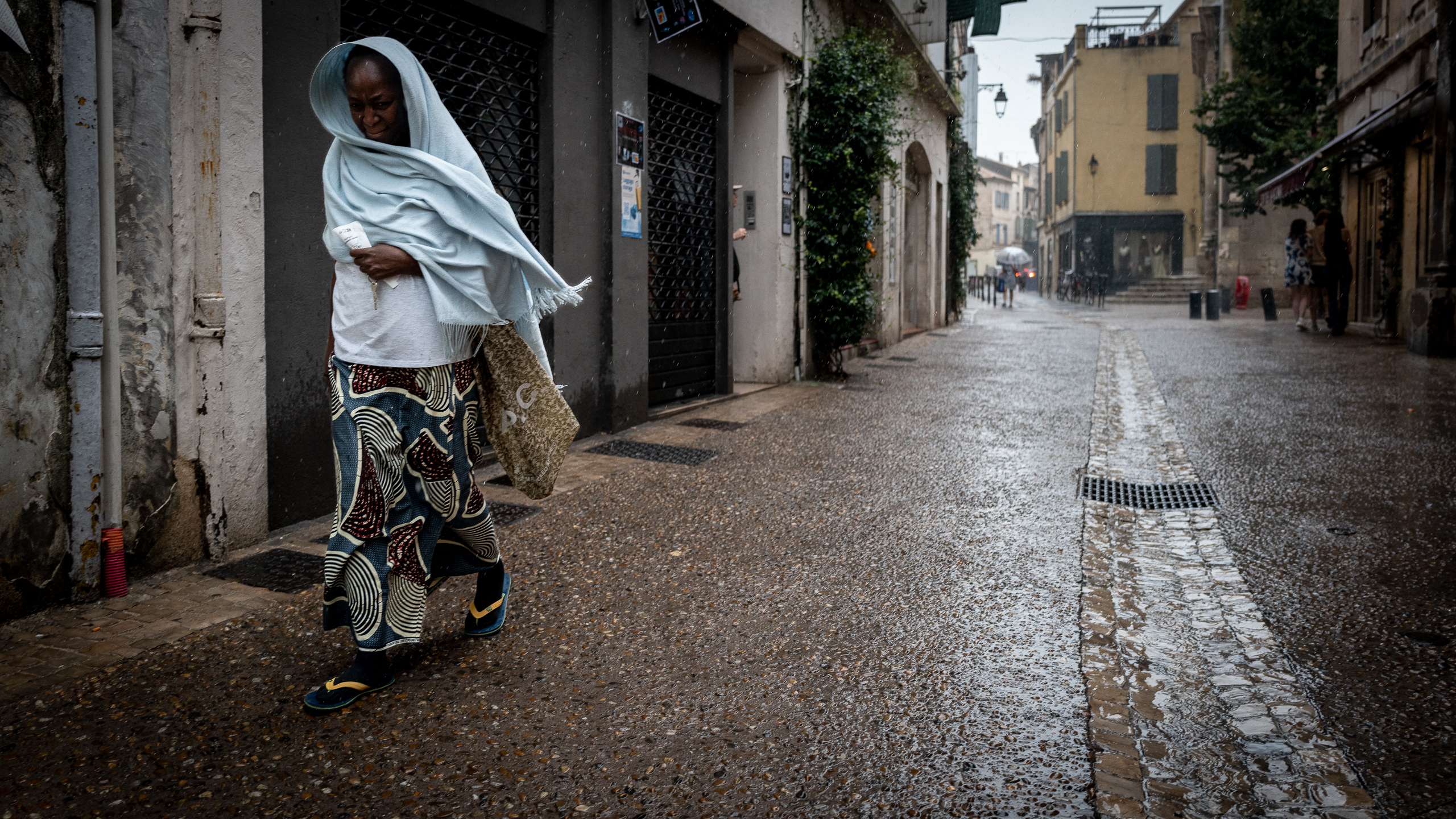 Femme marchant sous la pluie dans une rue, enveloppée d’un châle bleu clair